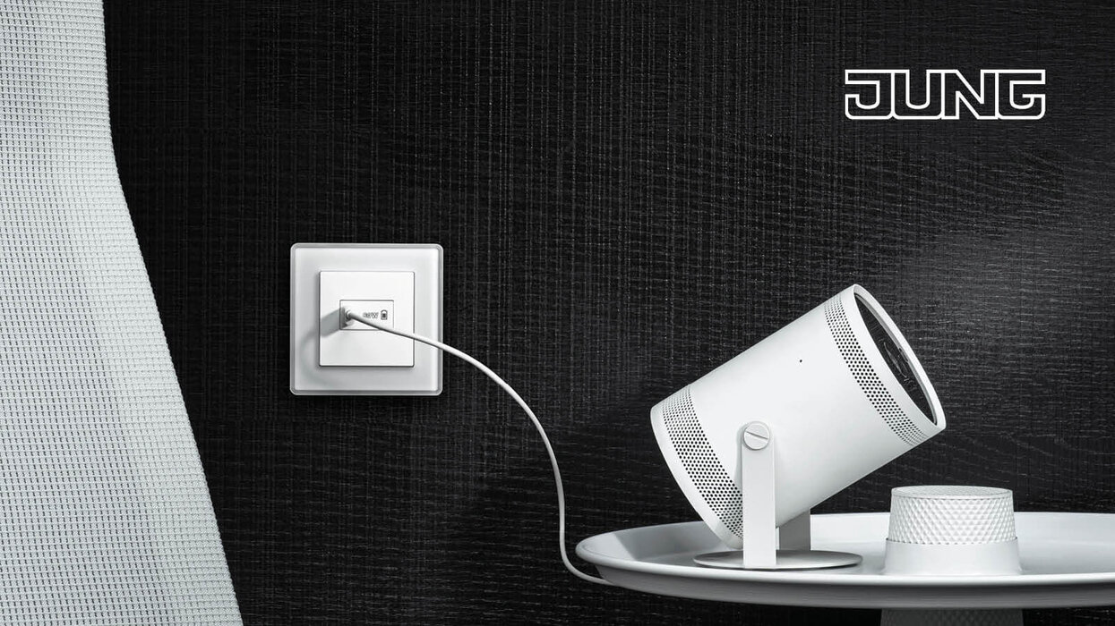 USB-Steckdose in der Farbe Weiß an einer schwarzen Wand. Es führt ein USB-Kabel zu einem Lichtspot, der auf dem Tisch steht.