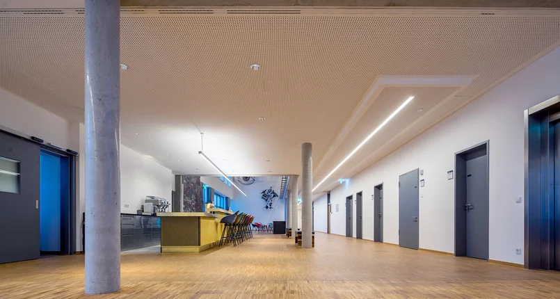 Das Bild zeigt einen großen Raum. An der rechten Raumseite gibt fünf graue geschlossene Türen.. Auf der linken Seite des Raumes steht eine gelbe Bar mit Zapfanlage. Davor stehen Barhocker. Im Raum sind vereinzelt Sitzgruppen verteilt. In die Raumdecke sind verschiedene Leuchtentypen eingebaut.