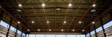 Das Bild zeigt eine große Halle mit Wellblechdach. Unter dem Dach ist ein Gestänge aus roten Metallverstrebungen befestigt. In regelmäßigen Abständen sind Hallenstrahler an Eckverbindungen der Verstrebungen angebracht.