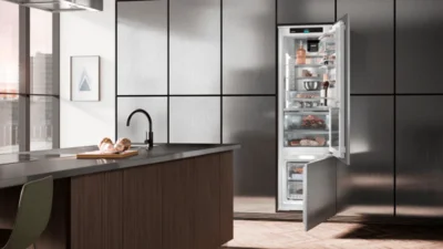 Das Bild zeigt eine moderne offene Küche. Im Vordergrund steht eine Kücheninsel mit Spüle. Eine Wand ist komplett mit silbernen Schranktüren bedeckt. In der Mitte der Schrankwand ist sind die Türen von einer eingebauten Kühl-Gefrierkombination geöffnet, so dass der jeweilige Innenraum erkennbar ist 