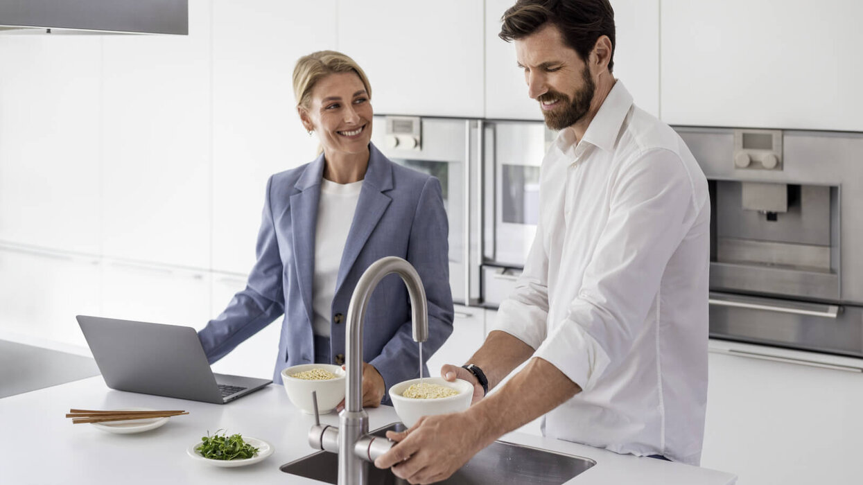 Zwei Personen, die an der Kücheninsel stehen. Eine Person bedient gerade den Wasserhahn, der in der Küchenzeile integriert ist.